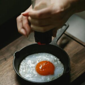 From above of crop anonymous female pouring savory from pepper mill on portion of fried egg in pan
